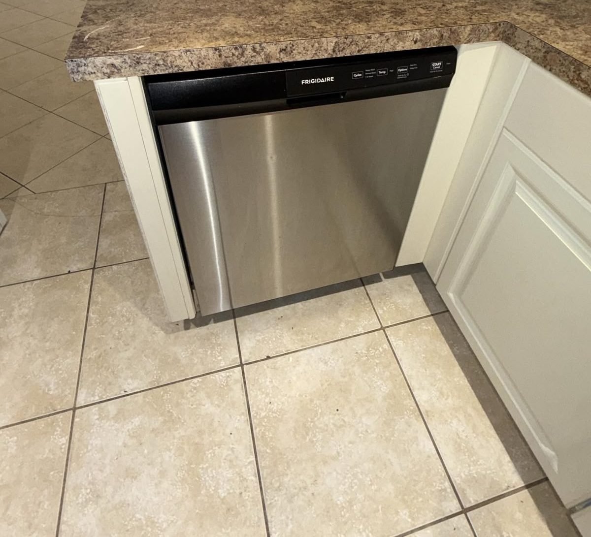 Stainless steel dishwasher front set into white cabinets, with the beige tile floor in front of it cleaned